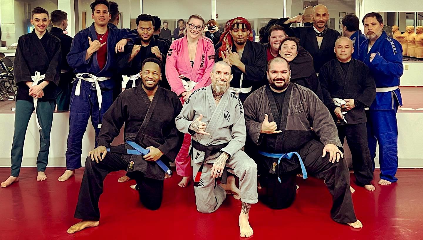 Group of diverse martial arts students and instructors at Amerikick Martial Arts in Northeast Philadelphia, showcasing camaraderie and various belt ranks, in a training environment with a red mat background.